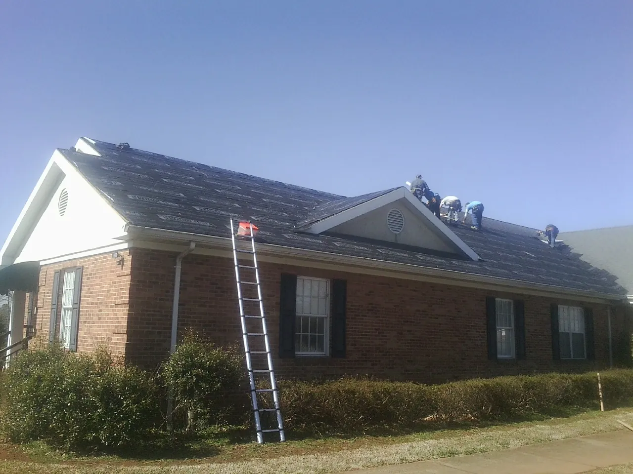 Crew tearing shingles off a brick ranch roof with a ladder set — Emergency Roof Repair work by our crew in Crossnore