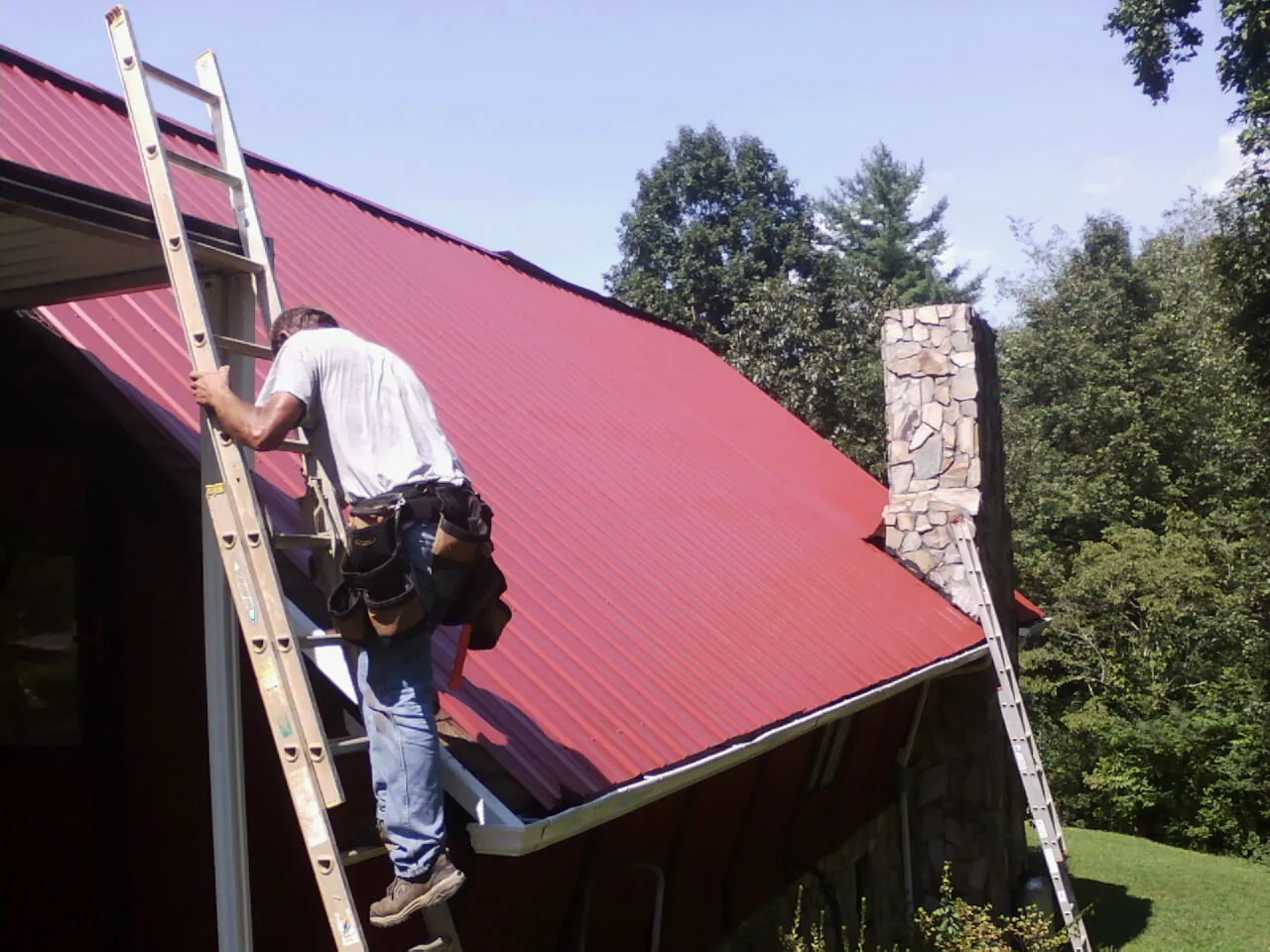 Roofer on a ladder beside a red metal roof and stone chimney — Soffit & Fascia Repair by Roofing Brothers in Crossnore